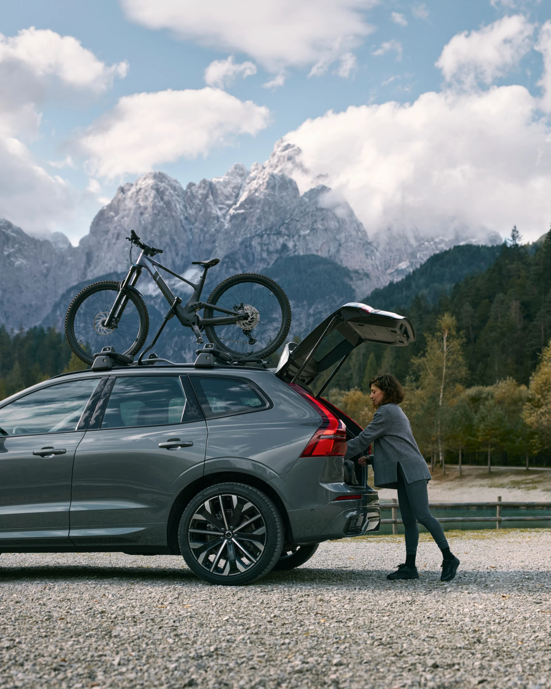 Женщина загружает снаряжение в открытый багажник Volvo XC60, припаркованного на живописной дороге, с велосипедом, прикрепленным к крыше.